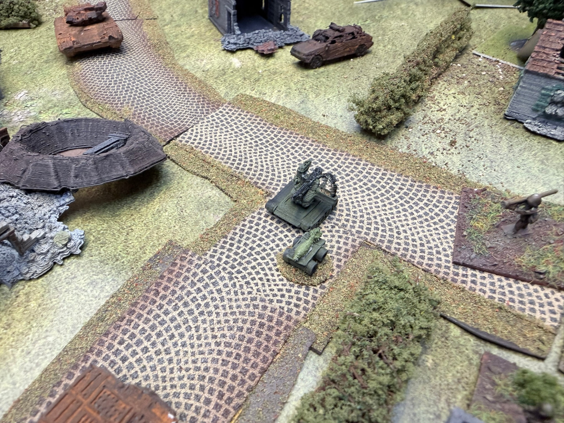 Two ground drones on a cobbled crossroads while an infantry miniature with a shoulder-fired rocket launcher watches